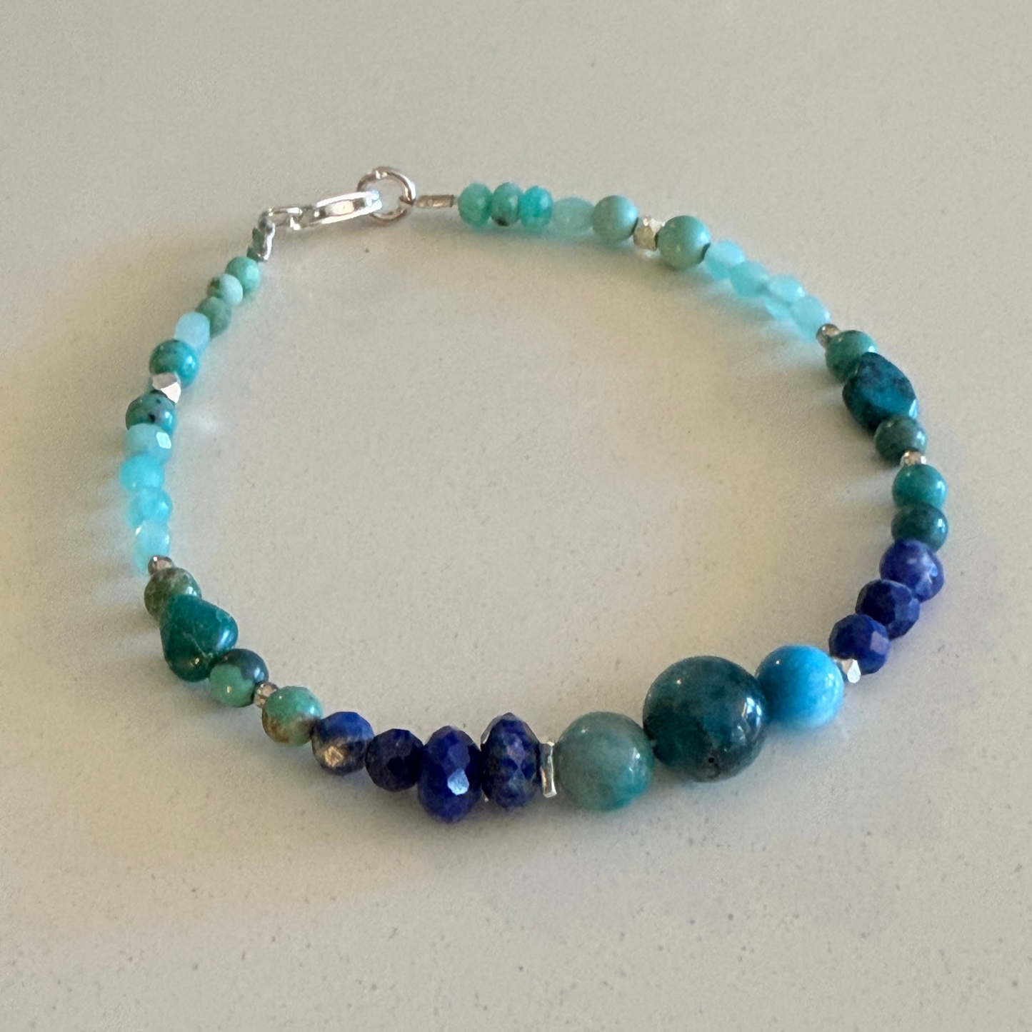 A beaded bracelet with a mix of turquoise, teal, and deep blue beads, accented by small silver spacer beads and a silver clasp, displayed on a neutral light beige surface.